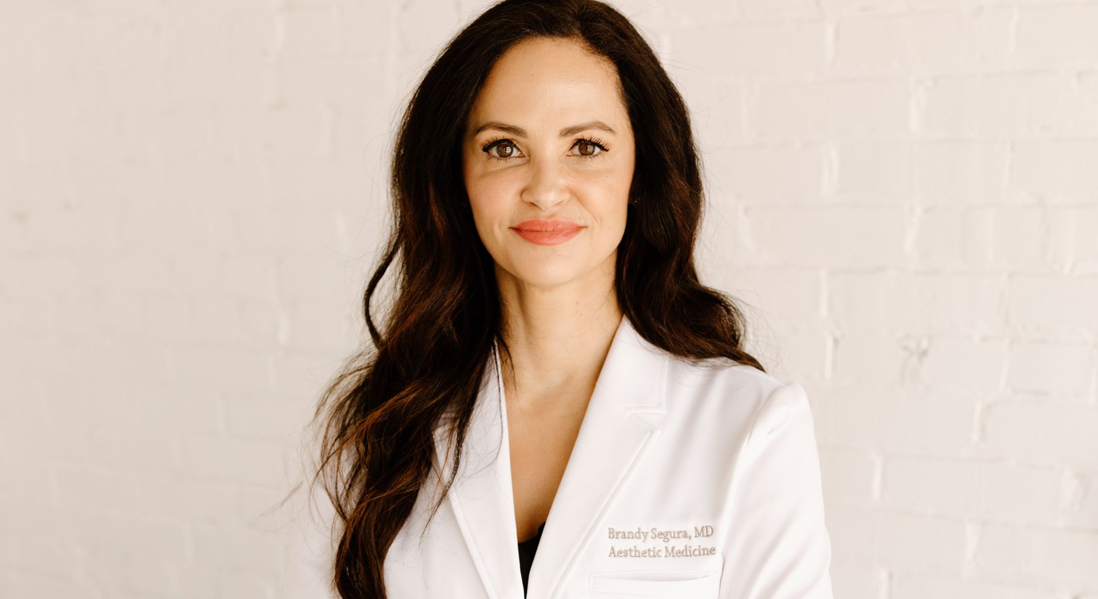 Doctor in white coat, smiling against white wall.